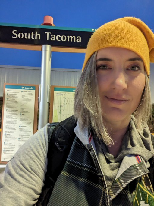 Sara Kiesler wears a yellow beanie and stands in from of the South Tacoma Sounder train station